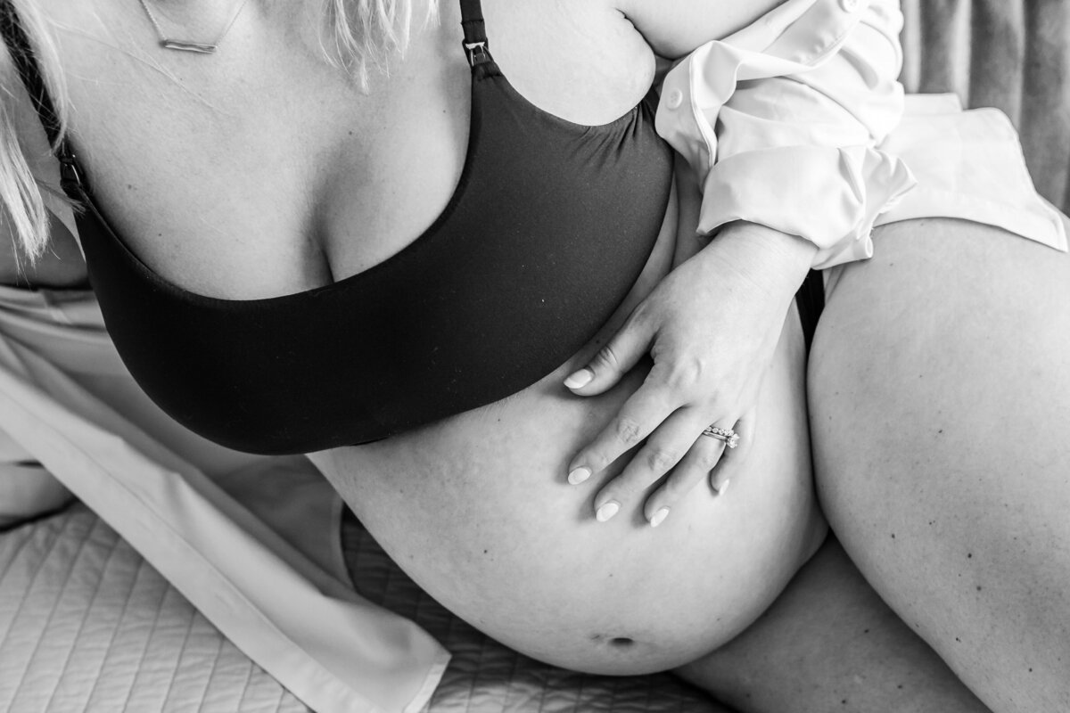A black-and-white close-up of a pregnant woman’s belly, with her hand resting gently on it, showcasing her engagement and wedding rings.