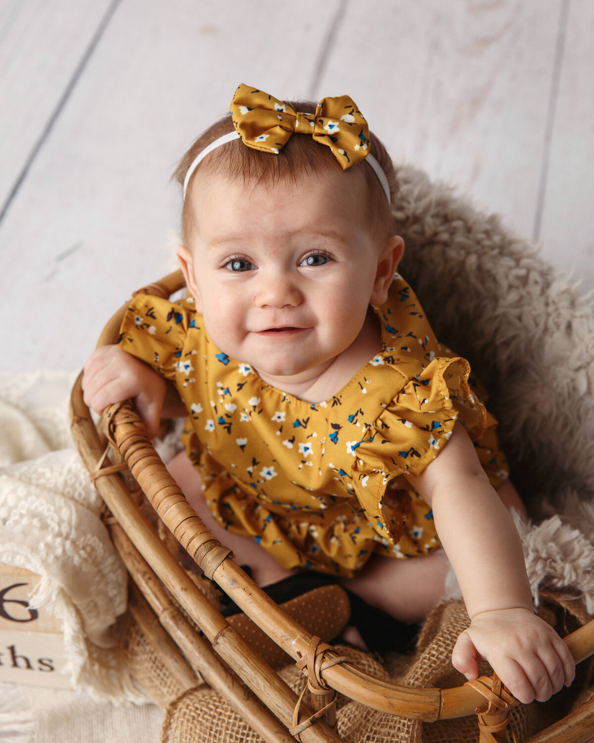 Cute baby girl in a yellow dress sitting in a wooden basket and smiling at the camera.