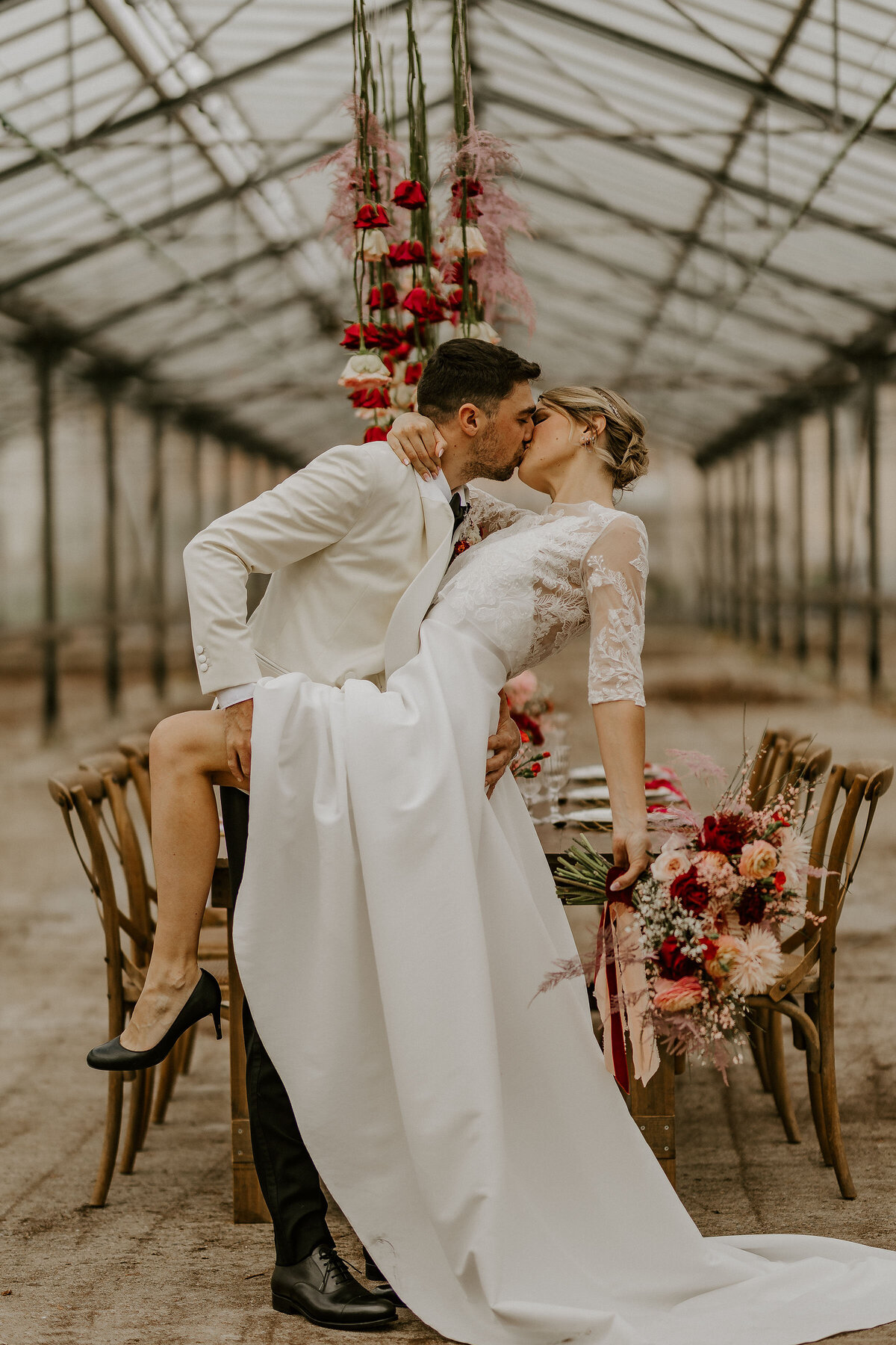 Couple s'embrassant dans un décor de mariage avec table et fleurs rouges et roses sous une verrière industrielle. Photo prise lors d'un workshop de photographie de mariage.