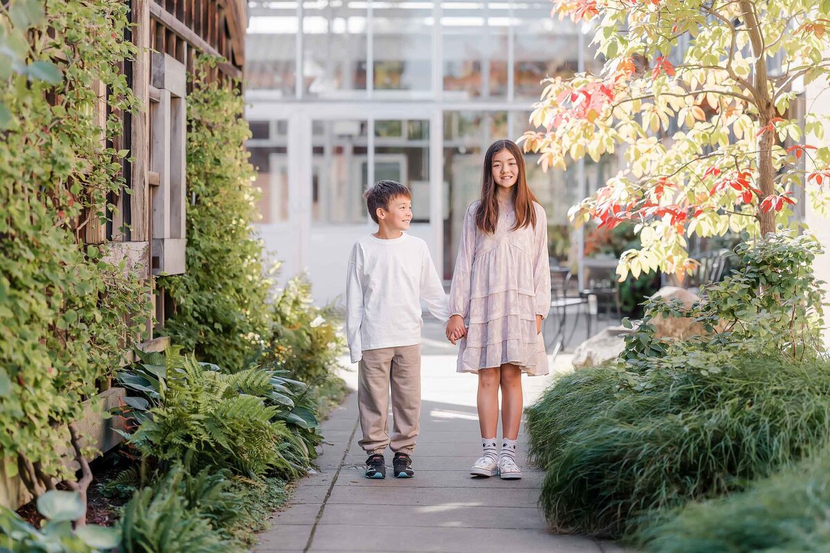 Classic portrait of a sister and brother holding hands at the Center for Urban Horticulture, surrounded by summer greenery.