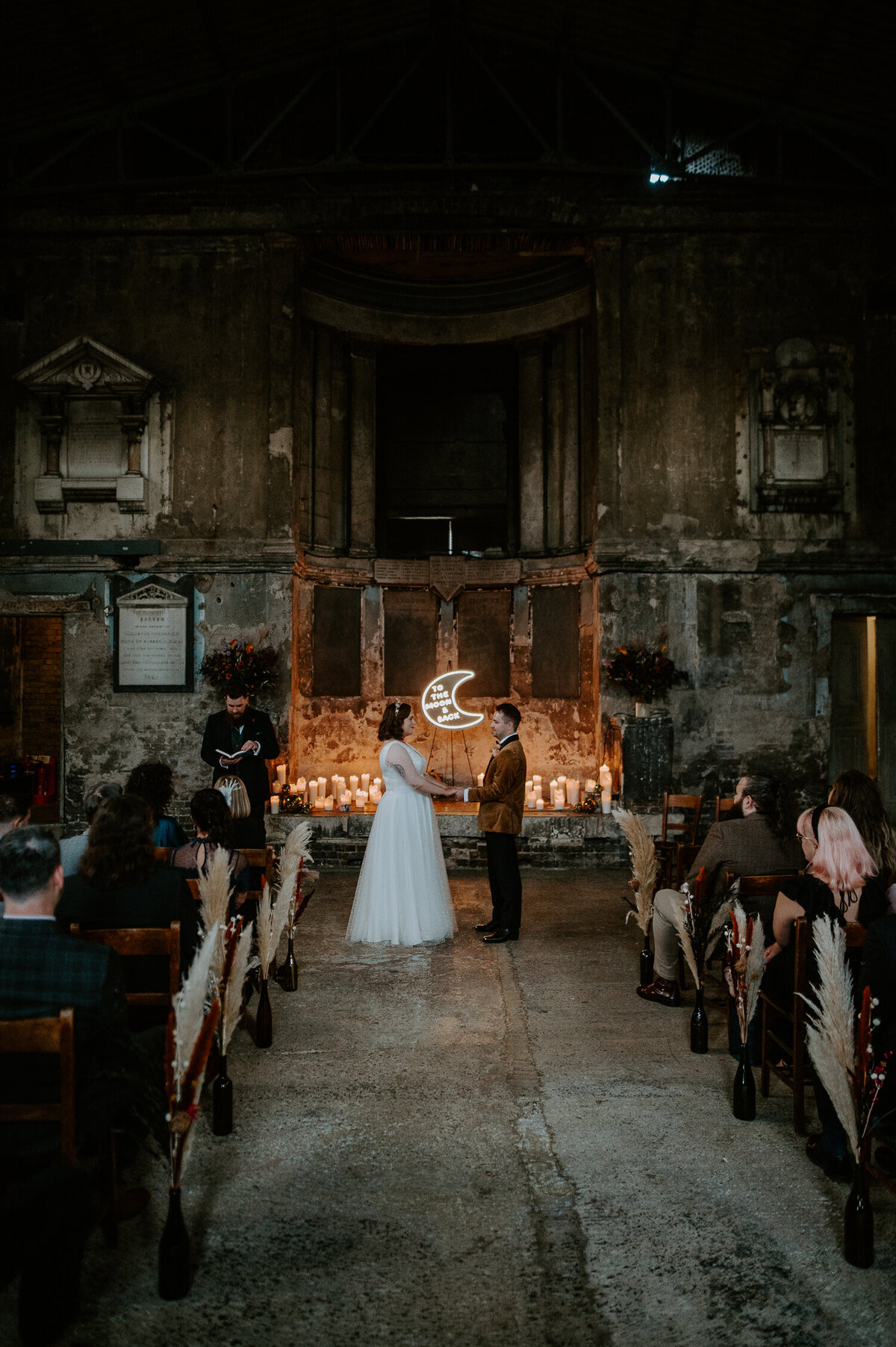 A wedding at the Asylum Chapel in Peckham London. There is a neon sign at the end of the isle that reads to the moon and back. A celebrant performs this very chilled ceremony whilst the couple are backlit by candles at the listed London venue.