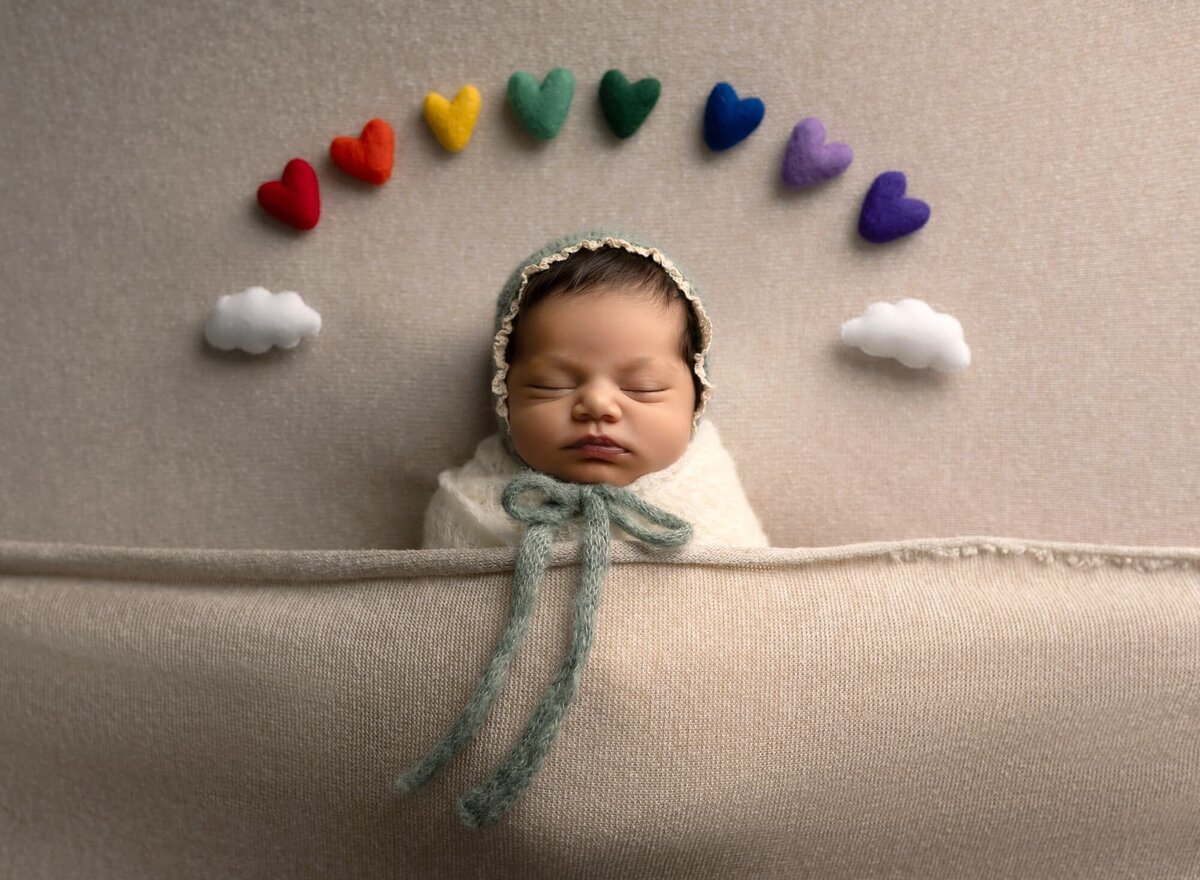 sleeping baby swaddled wearing green lacy bonnet with rainbow colored hearts over her head