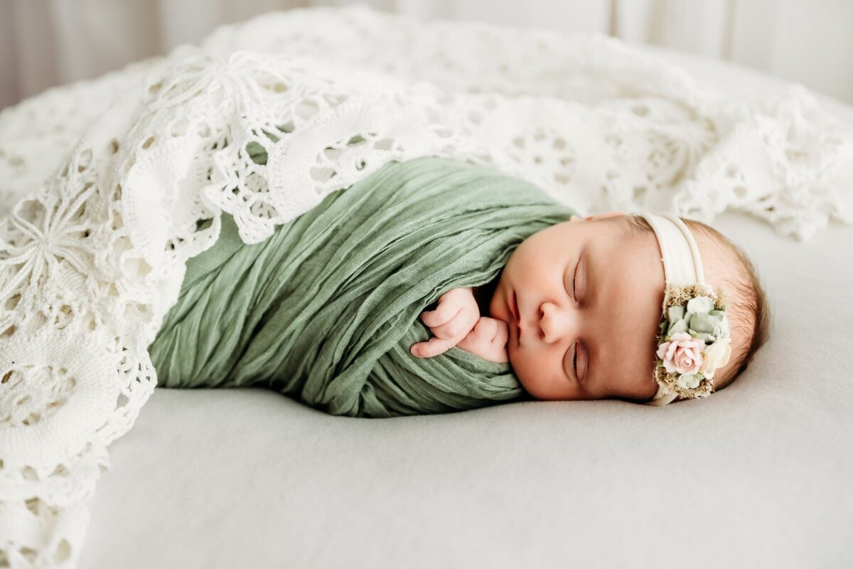 newborn swaddled in pale green. she is draped in a vintage lace and wears a green and pink flower on her head.