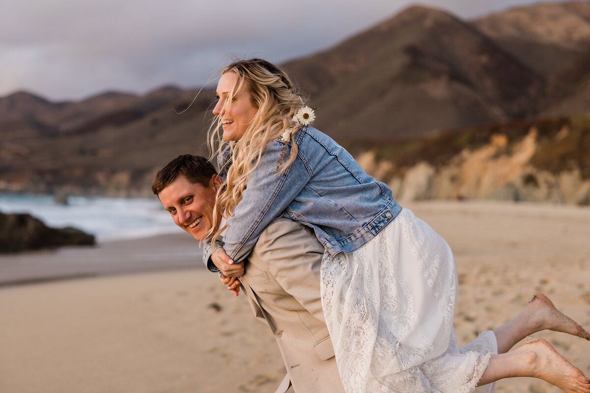 The groom gives the bride a piggy back ride on the beach in Big Sur