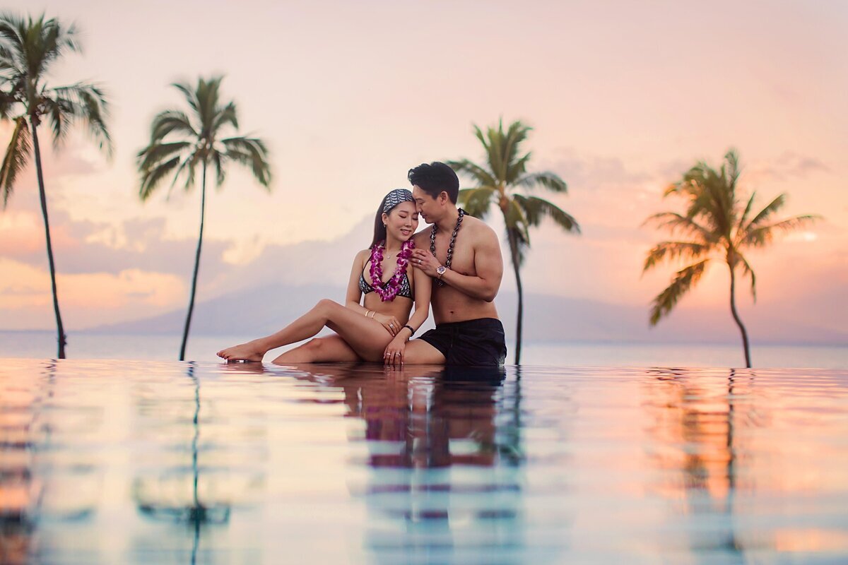 Engagement portraits taken at the Four Seasons Wailea infinity pool. Woman wears bikinis and a purple lei and husband kisses her cheek lovingly.