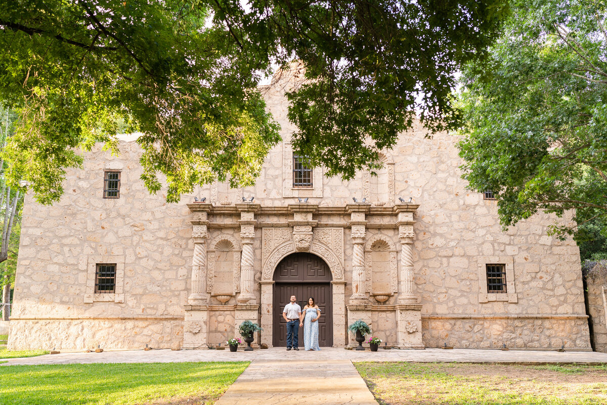 Husband and wife standing next to each other holding pregnant belly in front of Alamo structure at Cy Hope