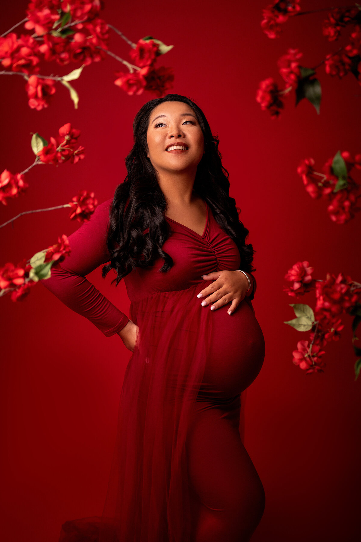 Pregnant woman in a deep red dress posing against a vivid red backdrop with floral accents, captured in a bold and modern maternity session in Waukesha.