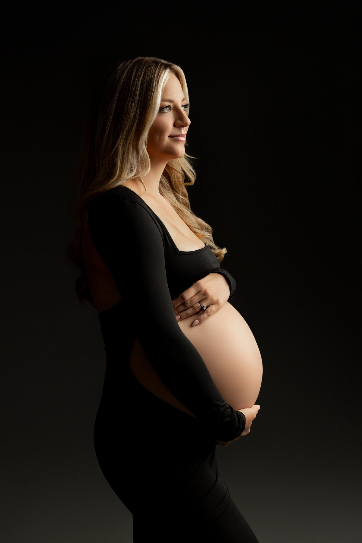 Stunning side profile of a glowing mother-to-be in a black form-fitting gown, showcasing her baby bump against a dark backdrop. Captured in a timeless maternity portrait by Inland Empire maternity photographer Kaitlyn Dawn Photography.