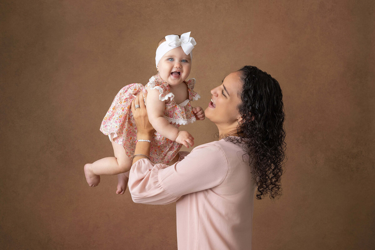 A woman with curly hair, wearing a light pink blouse, lifts a smiling baby girl dressed in a floral dress and a white bow headband. They stand against a plain brown background. Captured by a Jacksonville Family photographer, the baby's open mouth radiates joy.