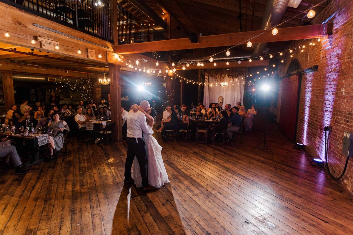 Bride and groom, sharing first dance in front of tables, full of cheering guests, with bright lights, spotlighting them