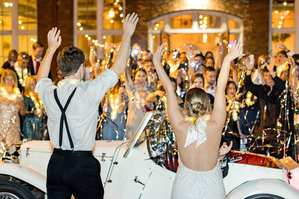 Golden hour wedding send-off filled with joy and celebration! The newlyweds raise their hands in excitement as they make their grand exit, surrounded by cheering guests, shimmering streamers, and twinkling lights. A vintage white convertible adds a timeless touch to this unforgettable moment. Captured by South Carolina luxury wedding photographer Jessica Hunt Photography.