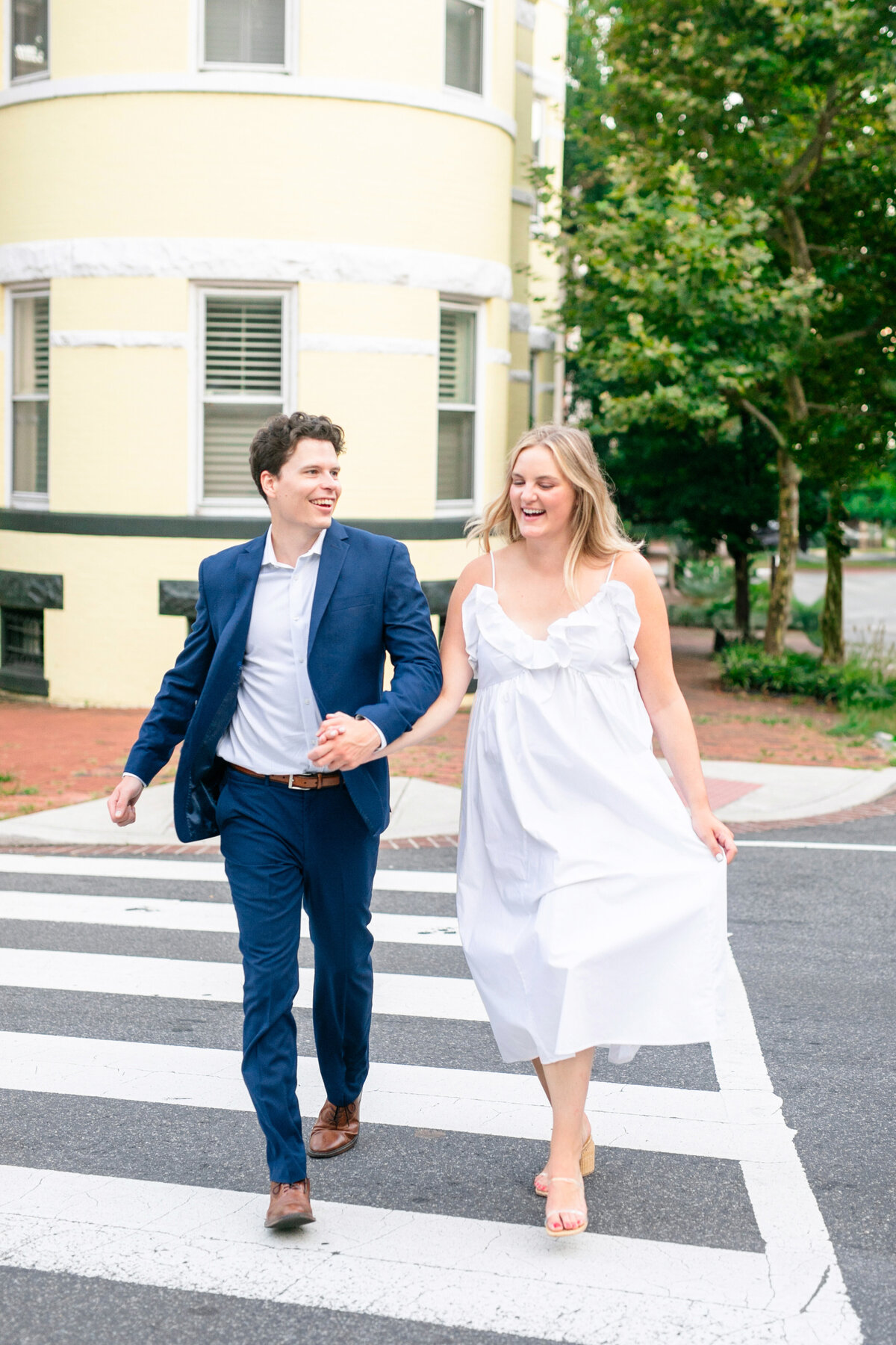 A couple joyfully walks hand-in-hand across a crosswalk. The man is wearing a blue suit, and the woman is in a flowing white dress. They are both smiling, with a yellow building and green trees in the background.
