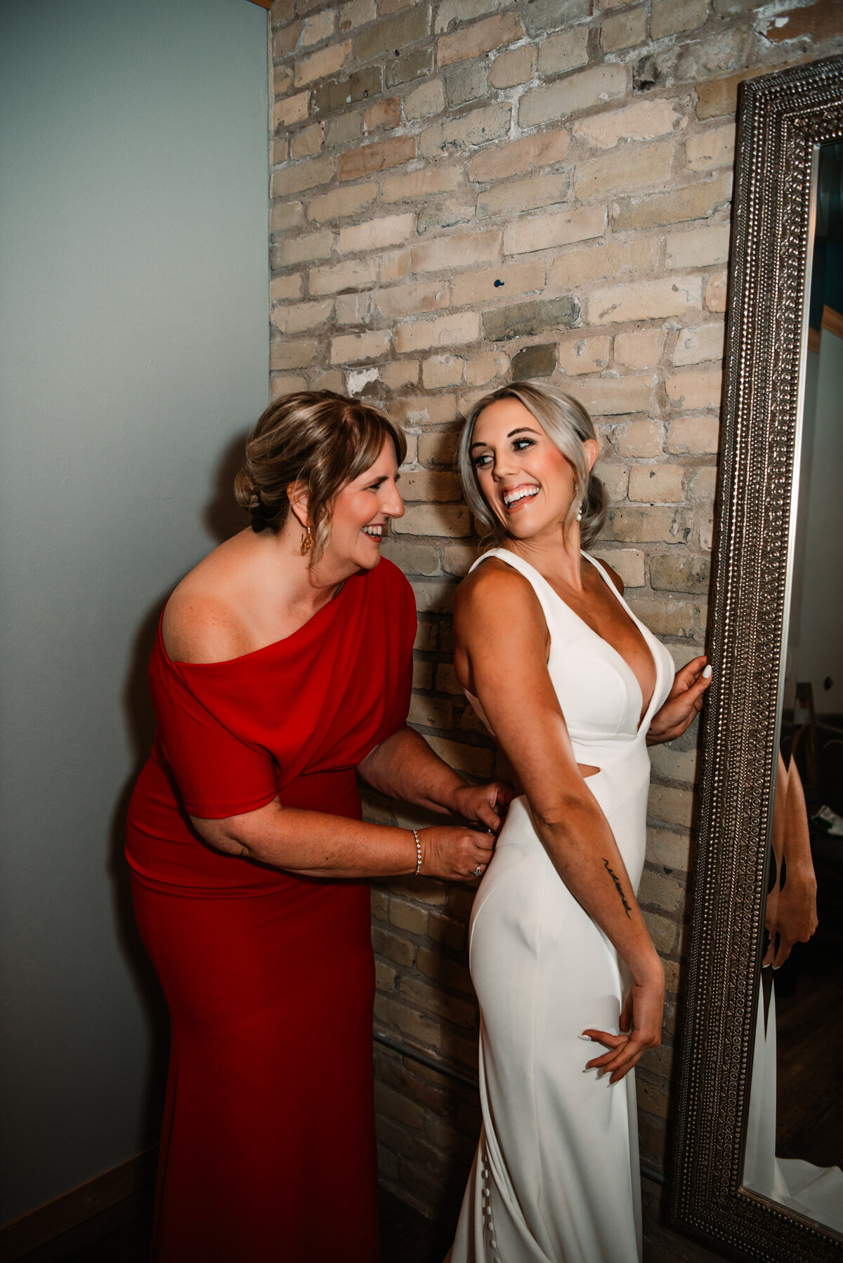 mom in red dress helps bride zip up her wedding dress