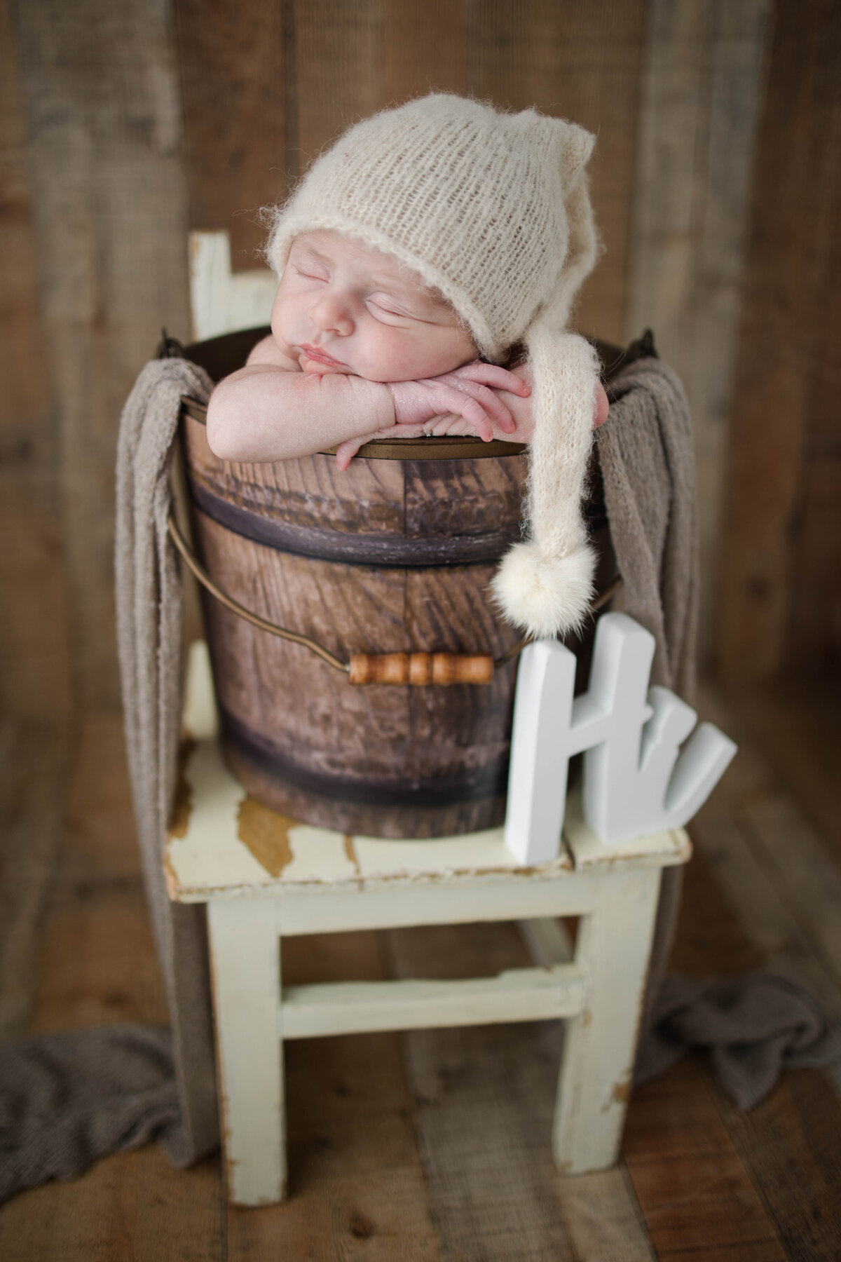Sleeping newborn baby in a wooden bucket with fluffy white hat and a hi sign