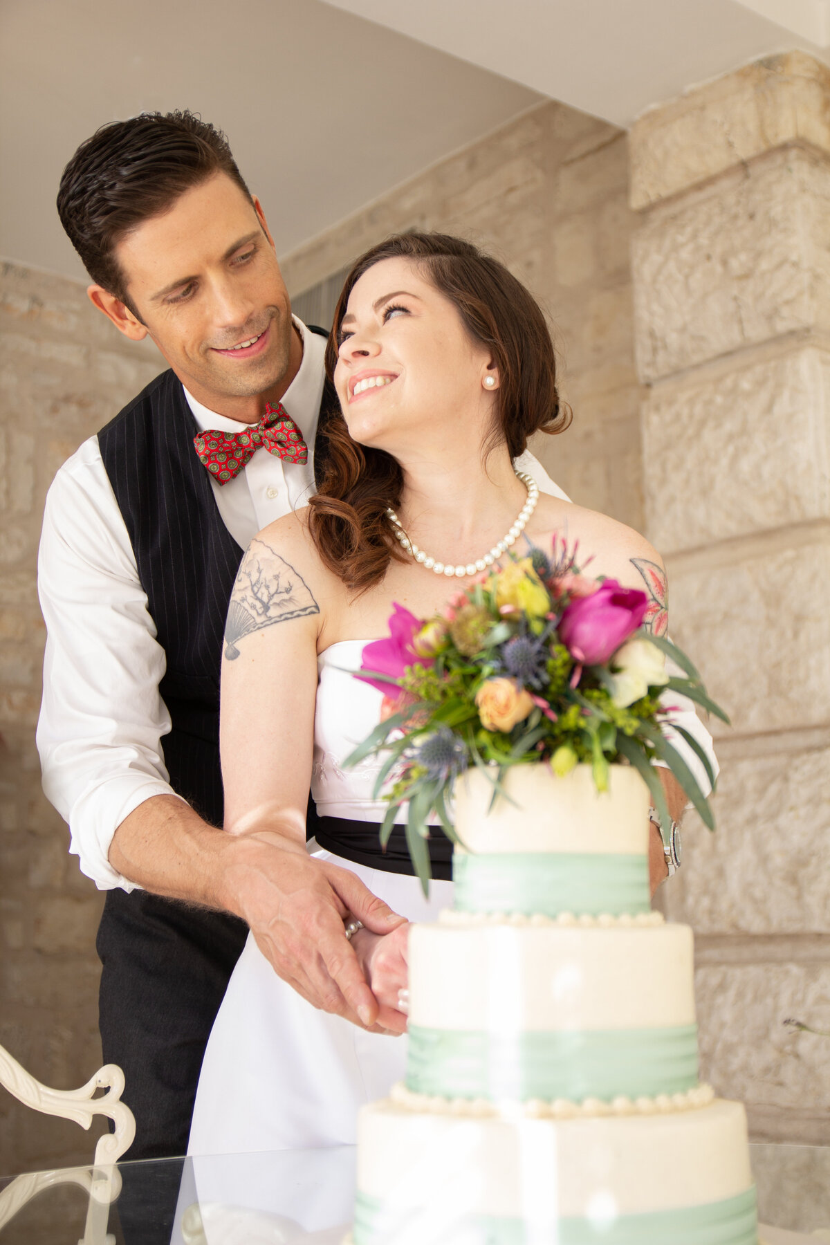 groom in vest and bow tie bride with tattoo on arm and pearls cut the cake at Chateau Bellevue  by Austin wedding photographer Firefly Photography