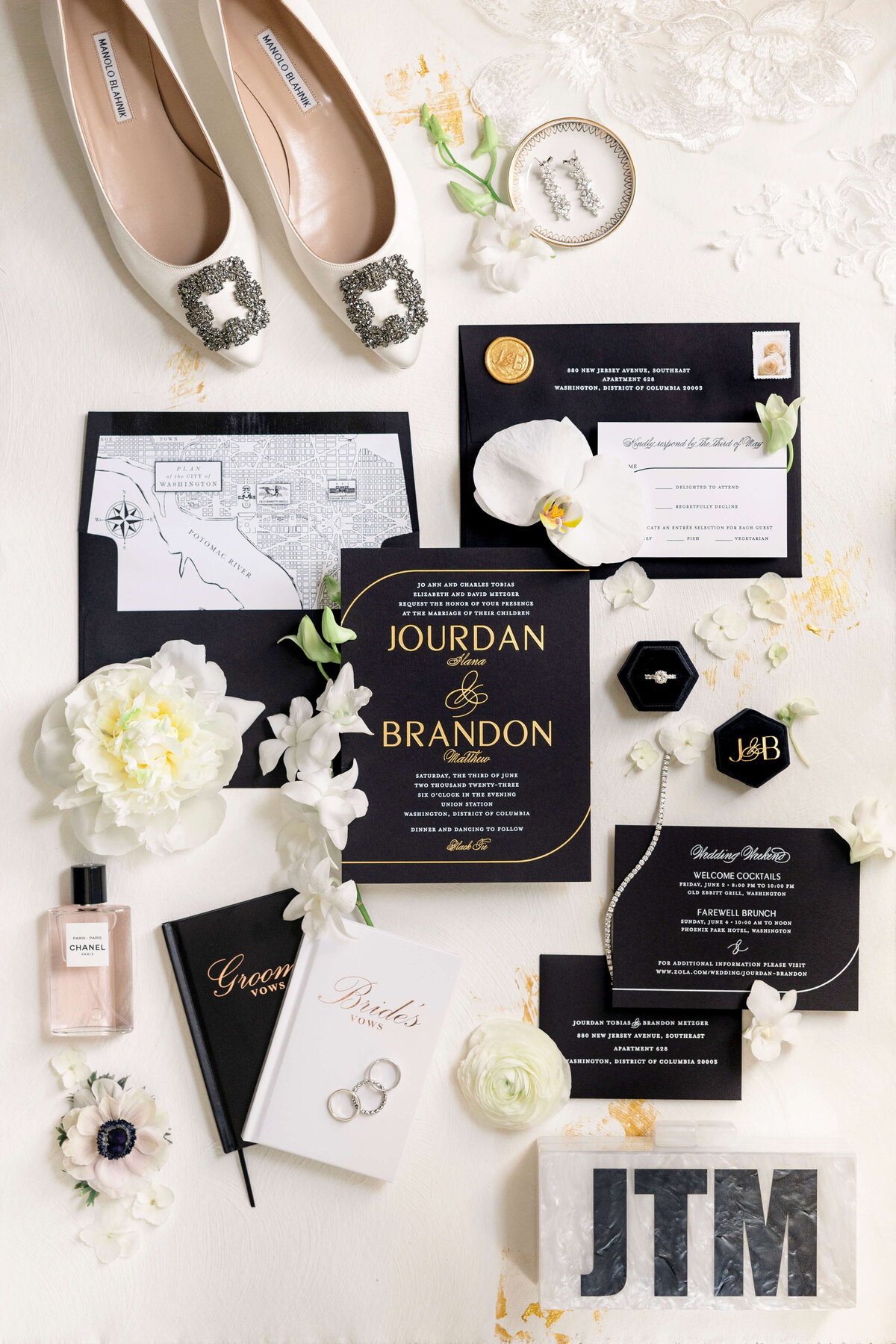Flat lay of wedding items including white shoes with jeweled buckles, black and white invitations, welcome notes, a floral map, rings, a perfume bottle, a small notebook, and decorative flowers on a light background.