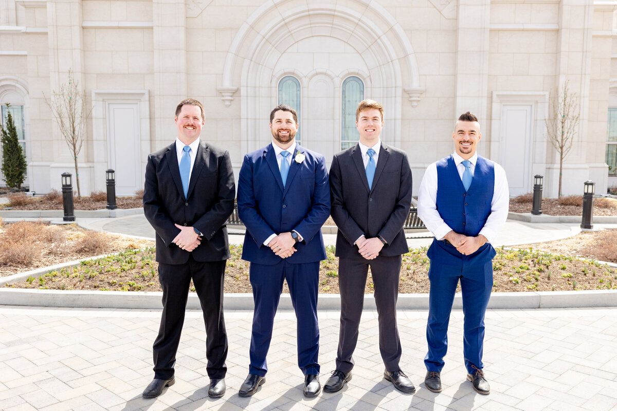 The groom stands with his groomsmen in a formal lineup, all wearing matching suits and blue ties in front of the temple.