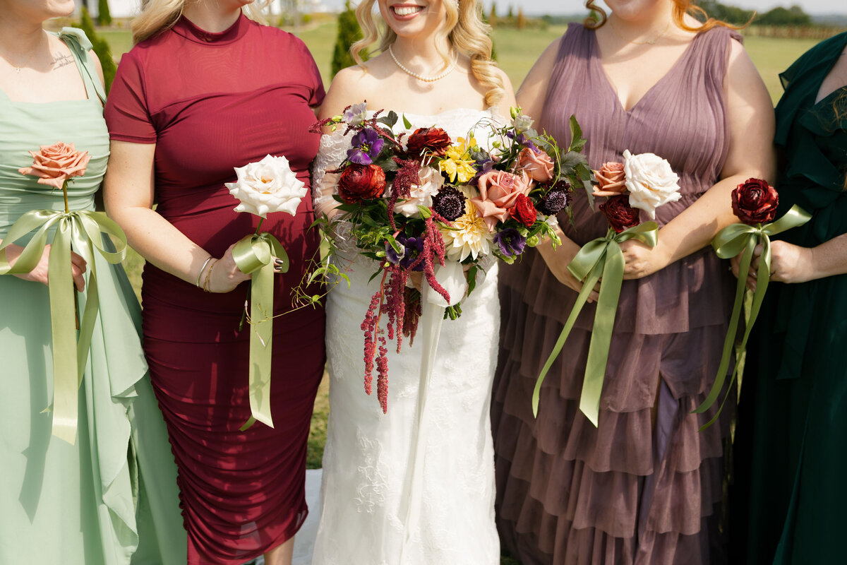 Close-up detail of the bride and bridesmaids holding their bouquets: the bride’s lush, colorful arrangement filled with dahlias, roses, scabiosa, and amaranthus, surrounded by bridesmaids holding single-stem roses tied with sage ribbon. A warm, romantic wedding color palette of burgundy, mauve, peach, and ivory.