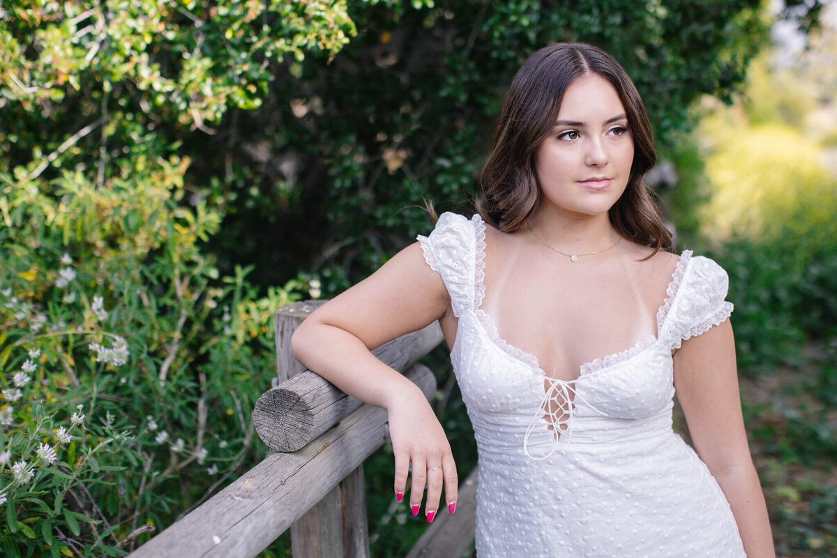 A young brunette woman wearing a white dress, resting her right elbow on a wooden park fence, looking away from the camera with greenery all around her. Captured at Northwest Open Space.