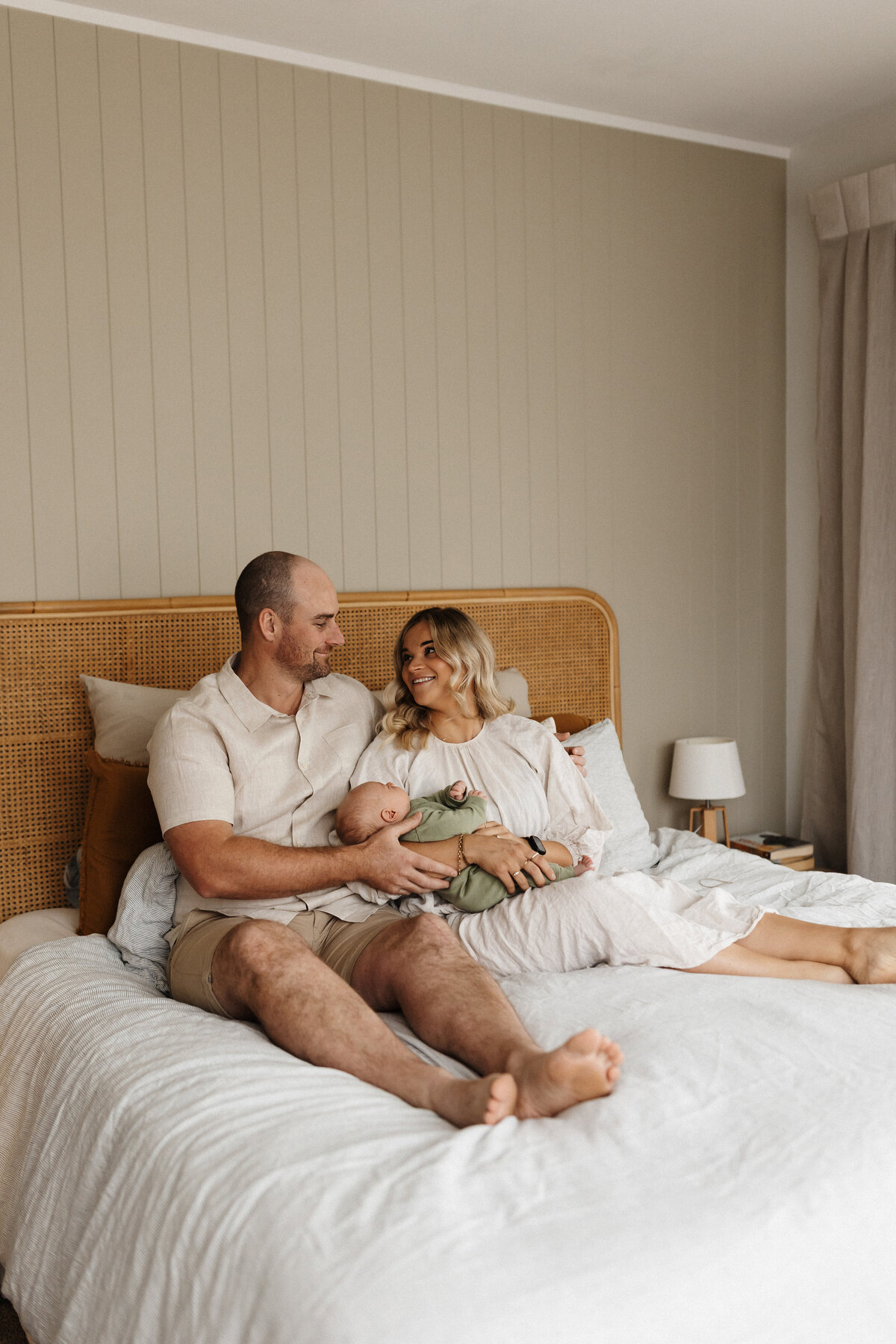 A mother and father sitting on their bed in their home holding their newborn baby while they're photographed by lucy jane photography during their christchurch in-home family photoshoot