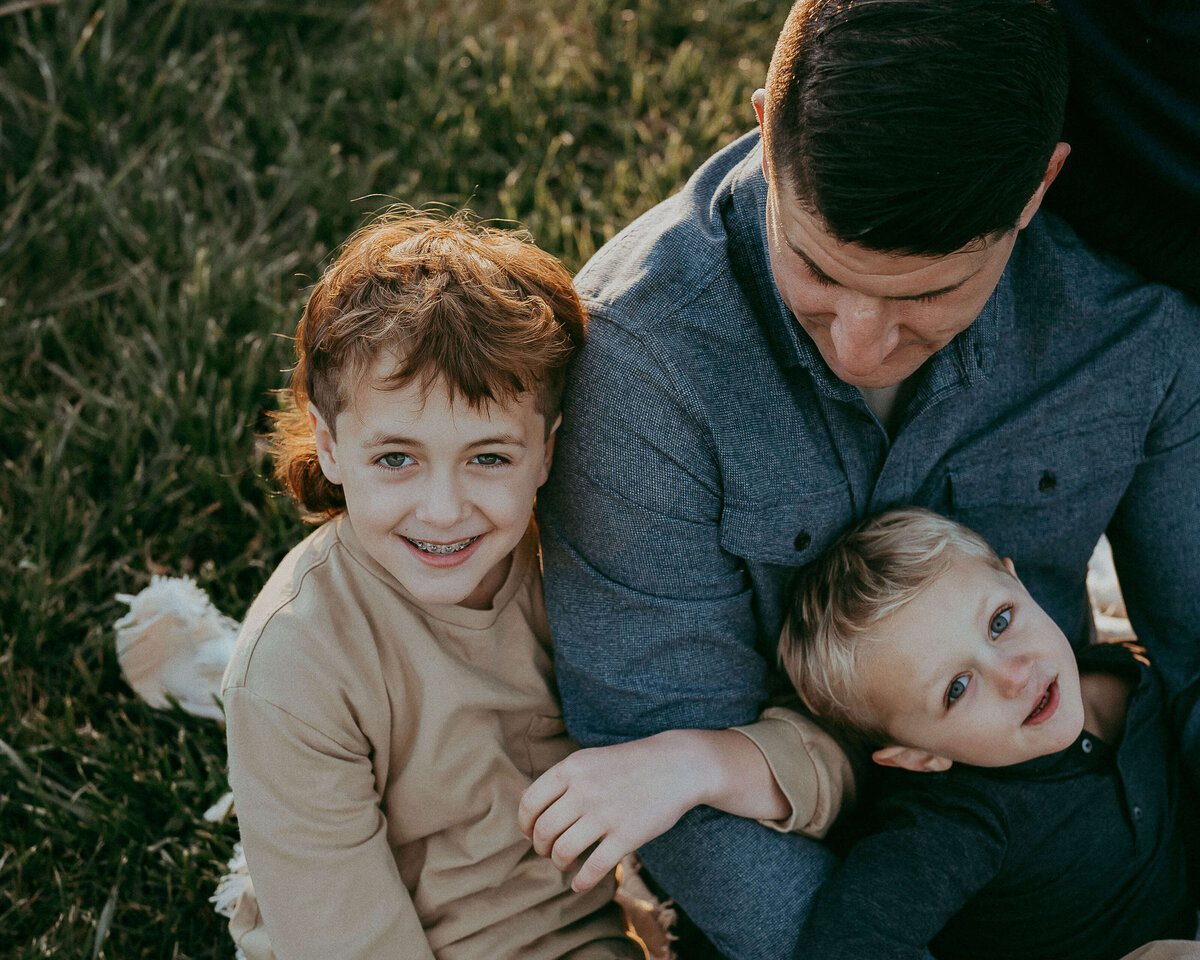 A boy wearing a tan shirt, hugs onto his dad's arm as the sun paints, a beautiful halo around their silhouette, his little brother lays across the dad's body, looking safe and content.