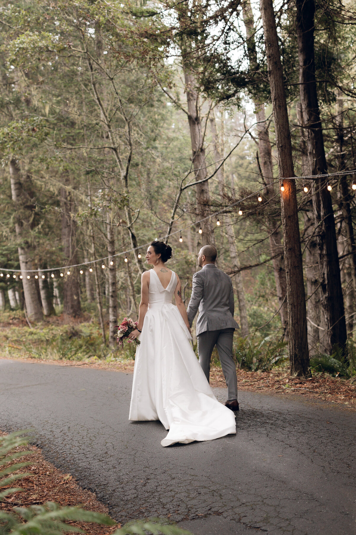 Cinematic wedding portrait featuring a bride and groom strolling along a forest-lined road, captured by a luxury wedding photographer emphasizing authentic storytelling and intimate moments.