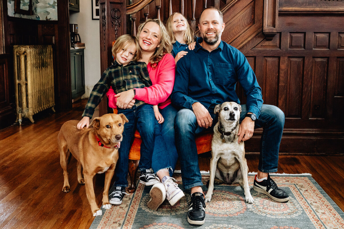 A family portrait indoors, with two children and two dogs. The parents sit close together, with the children by their sides, and the dogs at their feet, all in front of a wooden staircase.