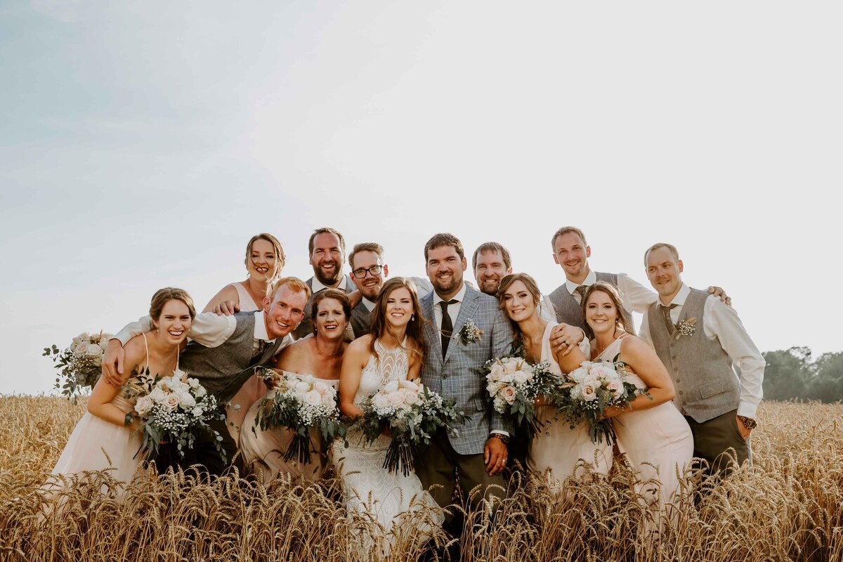 Bride, Groom, and wedding party are huddled together in golden wheat grass for rustic barn wedding in Exeter, Ontario.