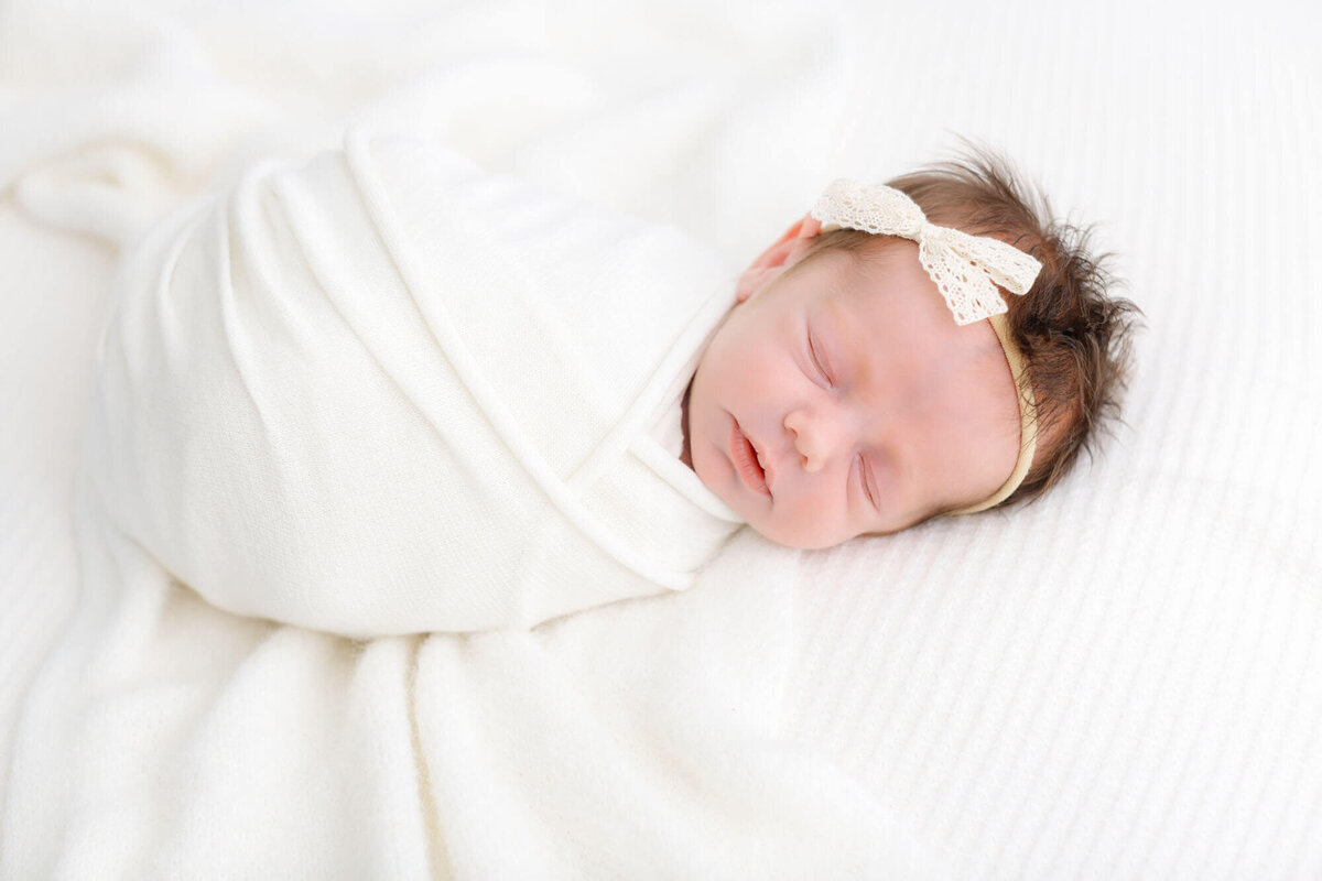 newborn baby wrapped in white and sleeping on side facing the camera. She is wearing a white neutral bow and is sleeping peacefully during her newborn photo session in Ashlie Behm Photography's Portland, OR studio.
