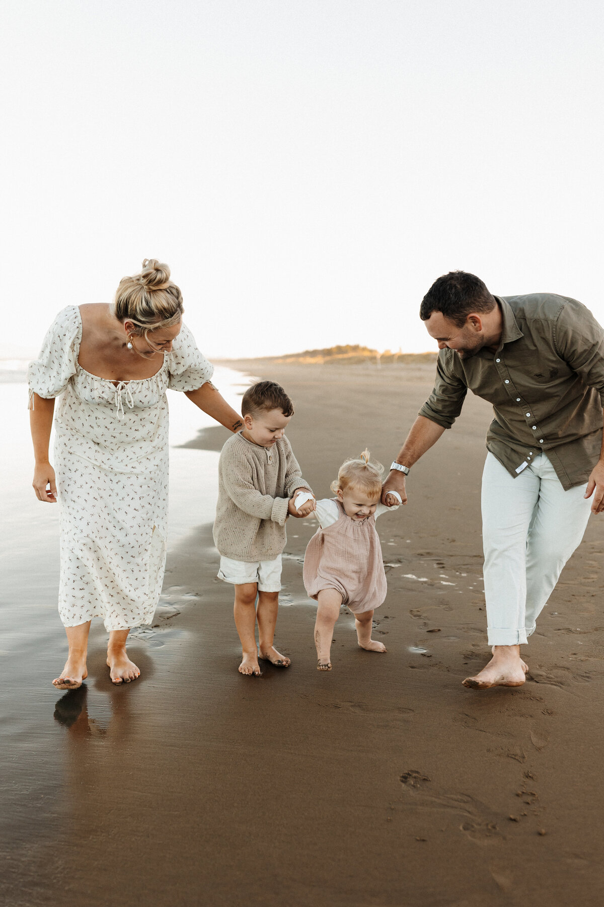 A mother, father and their two young children walking along the beach while the boy helps his little sister walking by holding her hands at the beach while lucy jane takes candid family photographs in canterbury