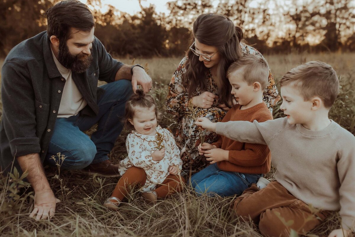 A family of five, including a man, woman, two boys, and a toddler, sits on grass in a field. The children are interacting, and the adults are watching them. Trees are visible in the background.
