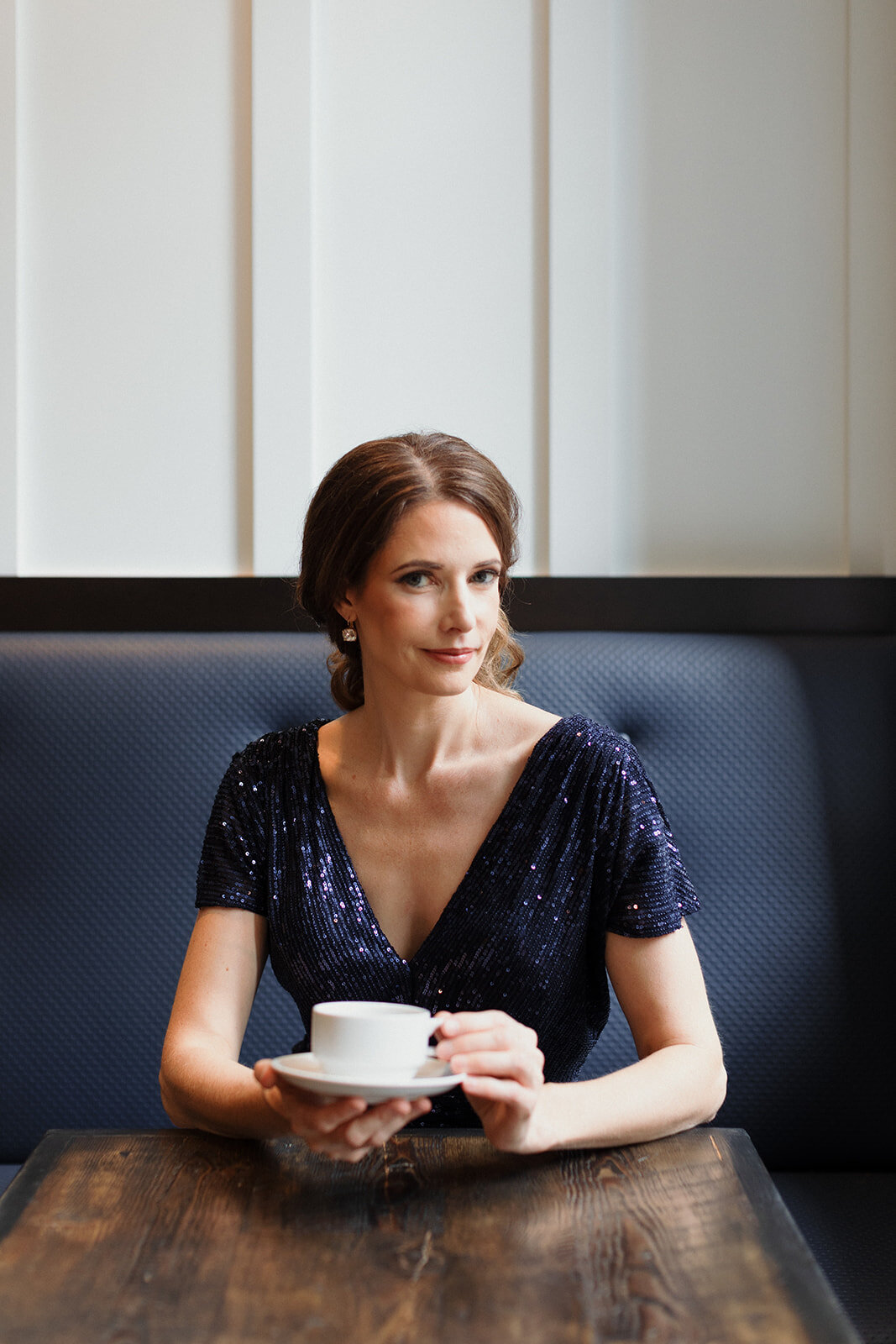 woman in dark blue sparkly gown is sitting in  restaurant booth, holding a coffee cup and looking at the camera with a knowing smile.