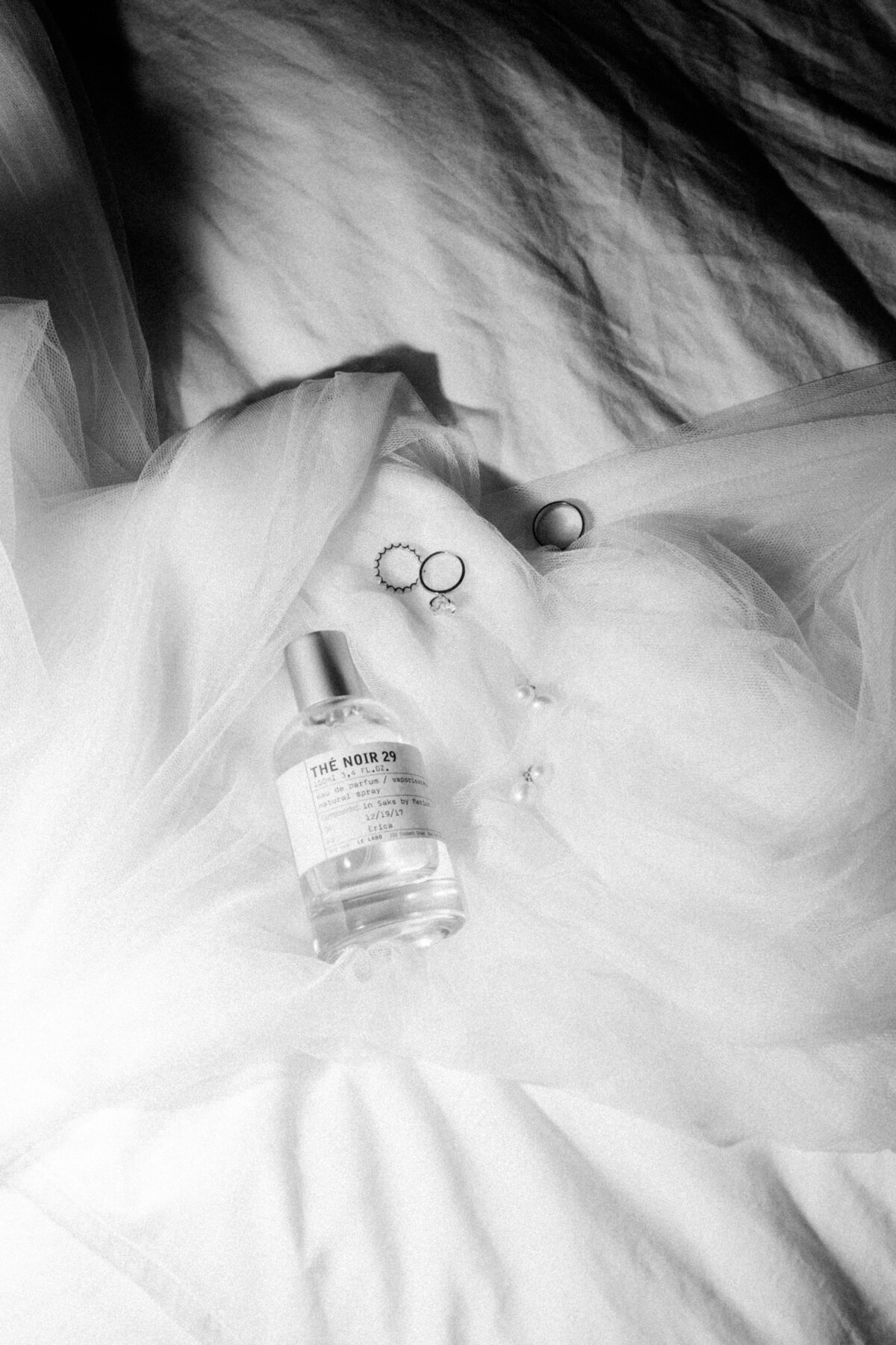 A beautiful still life of bridal accessories, including rings and a bottle of The Noir 29 perfume, delicately arranged on a soft, white veil for a maternity bridal portrait session