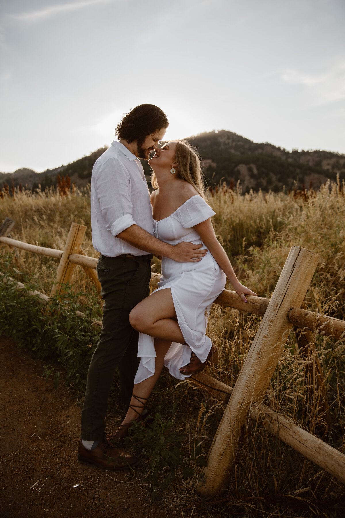 A couple leans in for a kiss at sunset, standing by a wooden fence in front of a mountain backdrop with golden grasses surrounding them.