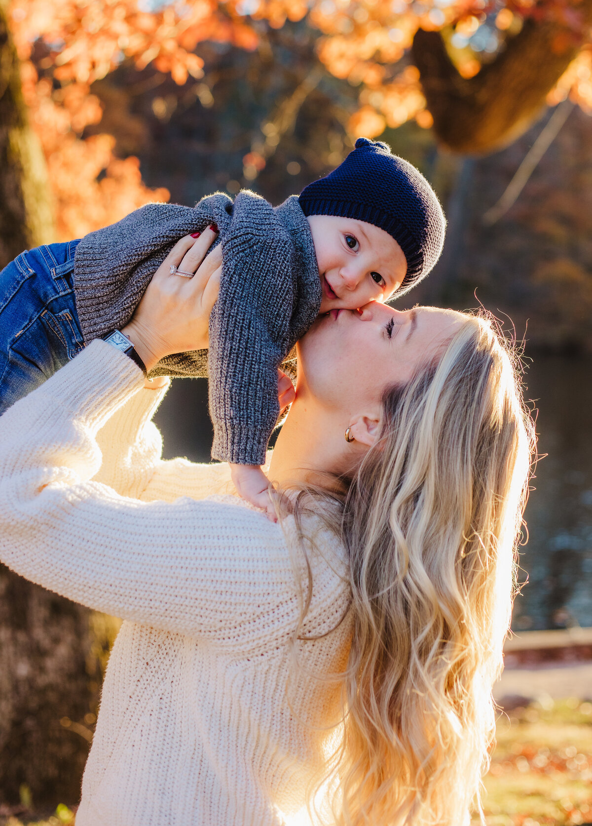 mother swinging baby boy up and kissing his cheek in front of autumn trees