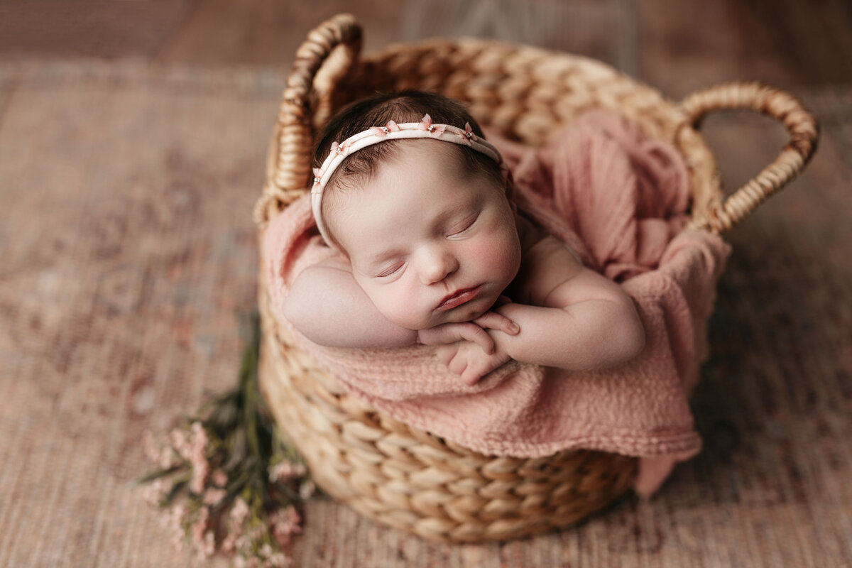 Studio newborn photography - Baby girl sleeping in a woven basket on a hardwood floor. Baby is resting on a pinky-apricot blanket with her hands folded under her chin.