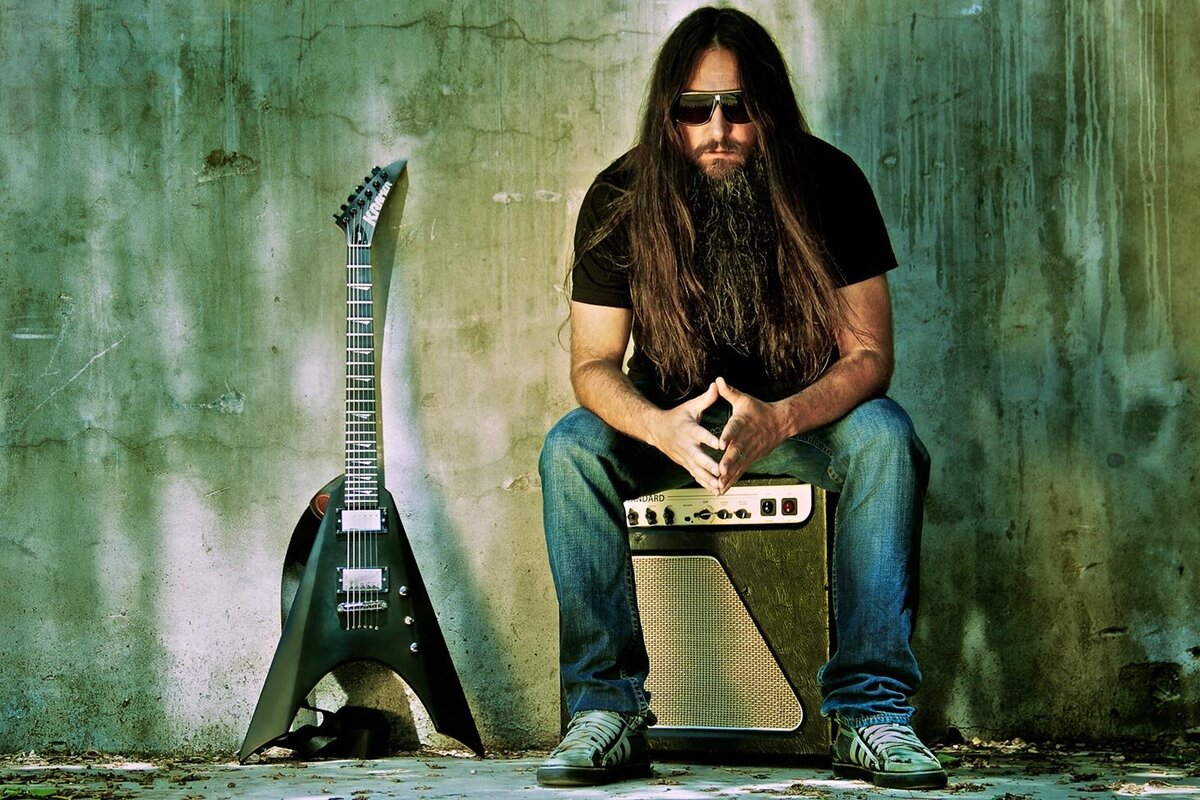 Rick Jacket musician portrait sitting on amplifier in front of green blue and yellow textured wall flying v guitar leaning against wall beside him