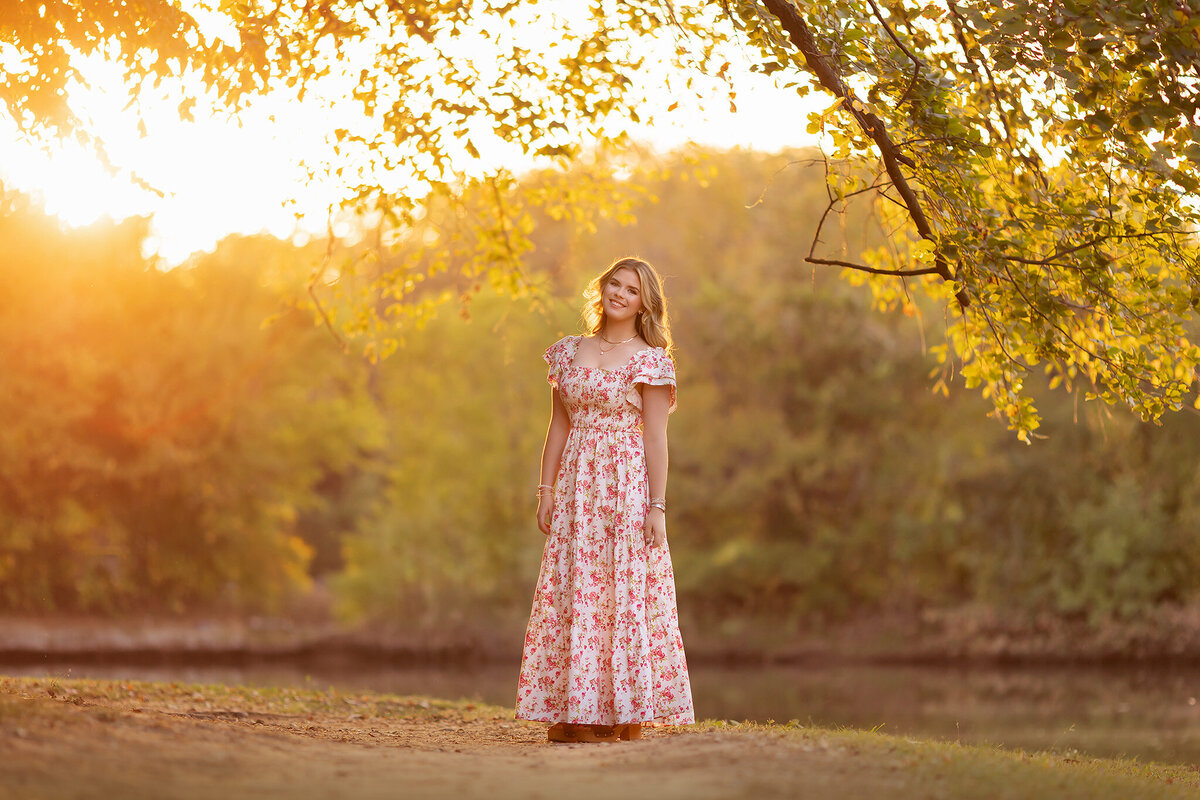 Dallas senior photography of a young woman in a floral dress, standing in golden hour light under a tree.