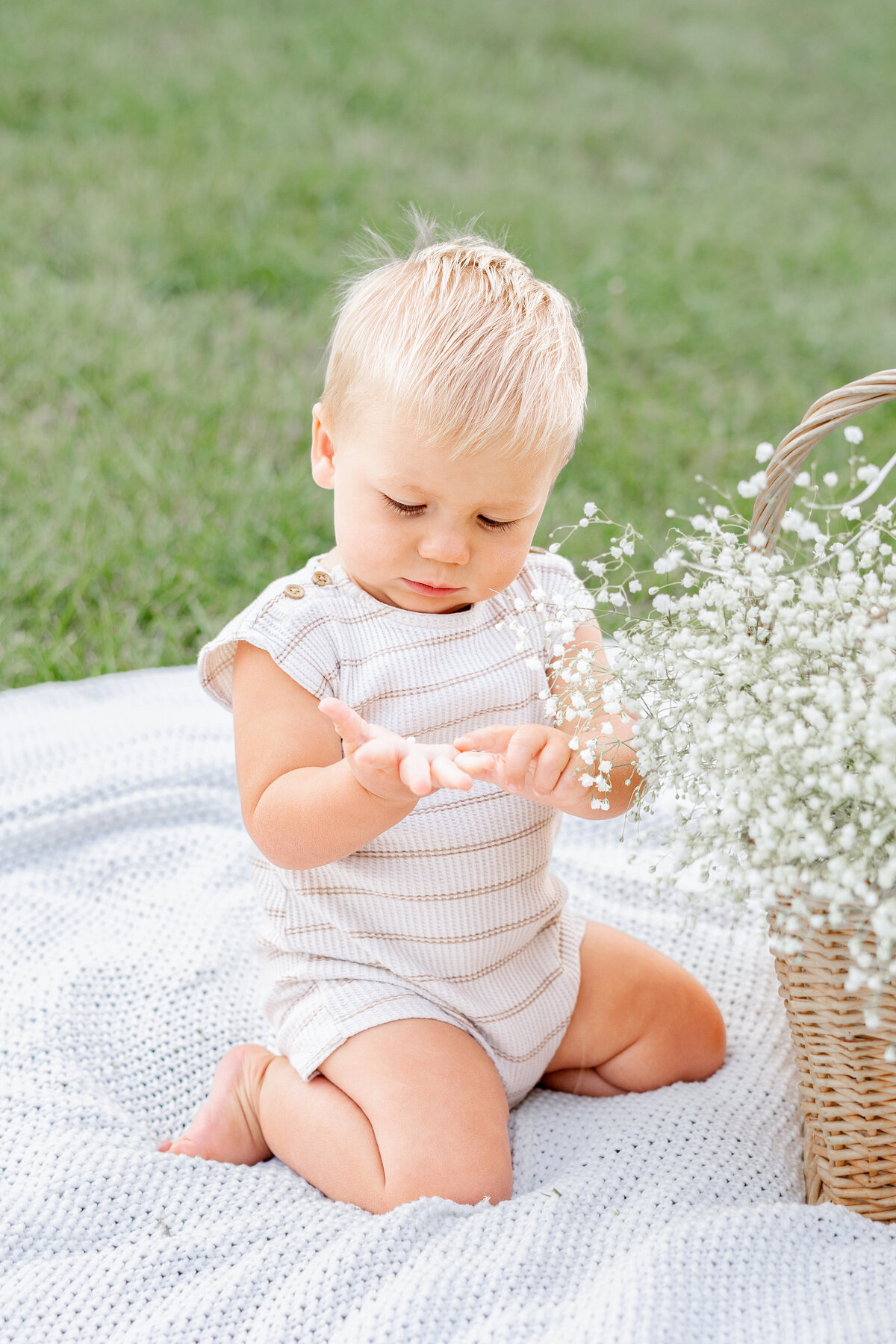 A blonde baby boy sits on a cozy blanket, fascinated by delicate white flowers in a woven basket, enjoying nature.