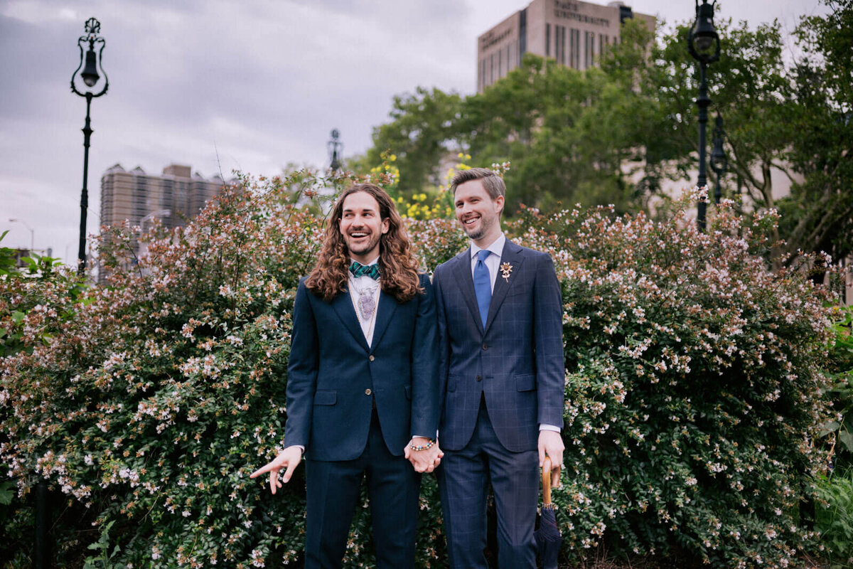 The two grooms are happily holding hands, with plants in the background. NYC City Hall Elopement Image by Jenny Fu Studio