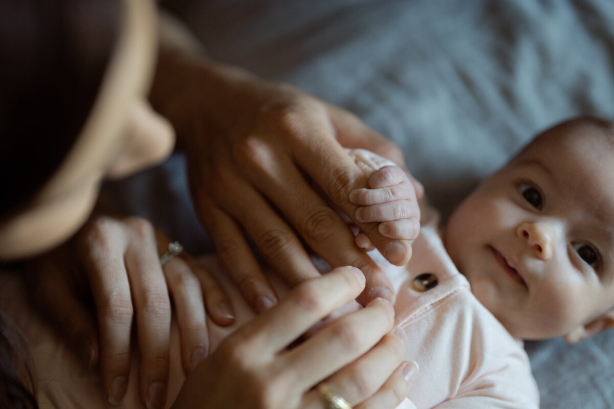 An endearing close-up of a baby grasping their parents' fingers while lying comfortably on a soft surface. The baby's eyes are full of curiosity, reflecting the warmth and connection of this tender moment. Captured during a lifestyle family session in the family's home and baby nursery in Fort Worth, Texas.