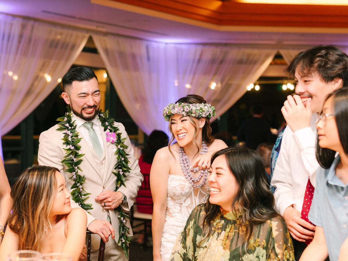 A joyful wedding gathering with guests smiling and laughing. The bride, in a floral crown and white dress, stands beside the groom in a beige suit with a lei.