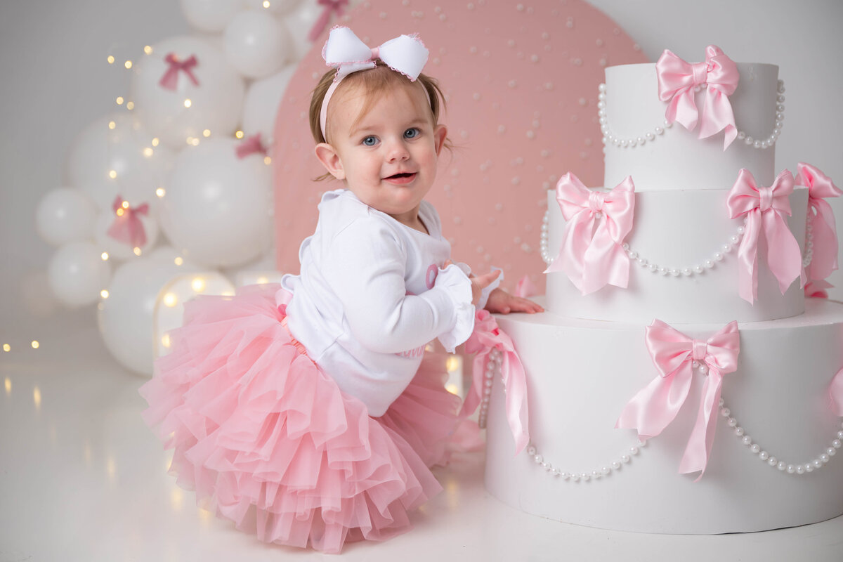 Close up of a child kneeling next to a cake prop decorated with pearls and bows.
