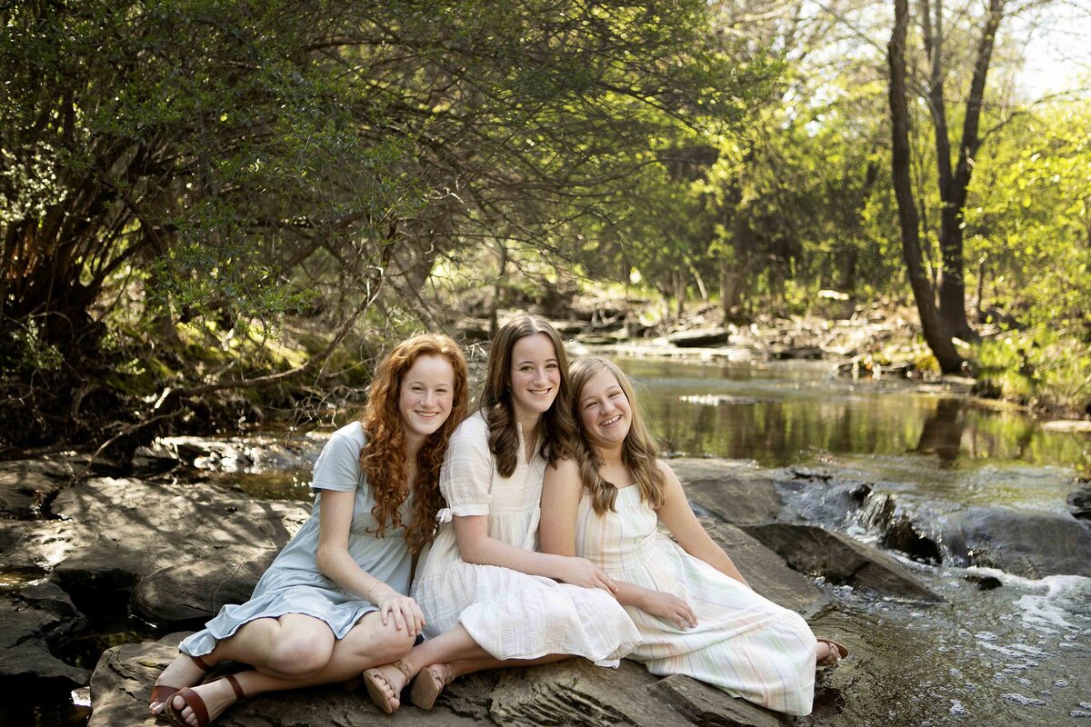 The three sisters sitting together by the creek in Flower Mound, enjoying a peaceful moment while posing for their photoshoot