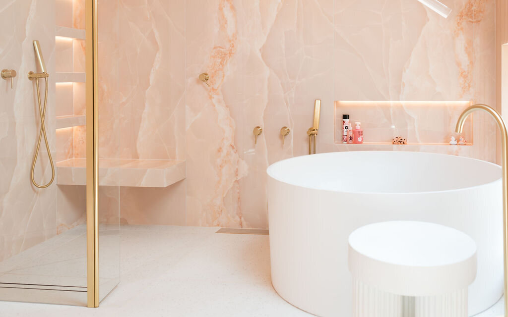 Luxurious bathroom featuring a freestanding round bathtub, gold fixtures, and pink marble walls. A glass-enclosed walk-in shower with built-in shelving is to the left. The room has a clean, modern aesthetic.