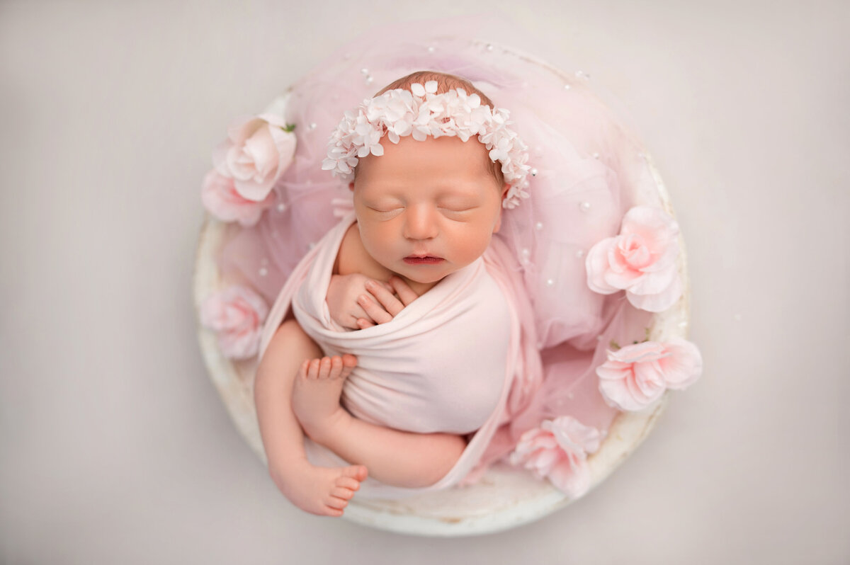 newborn baby girl wrapped in pink swaddle posing inside of white bowl surrounded by pink pearl tulle and pink flowers on cream backdrop