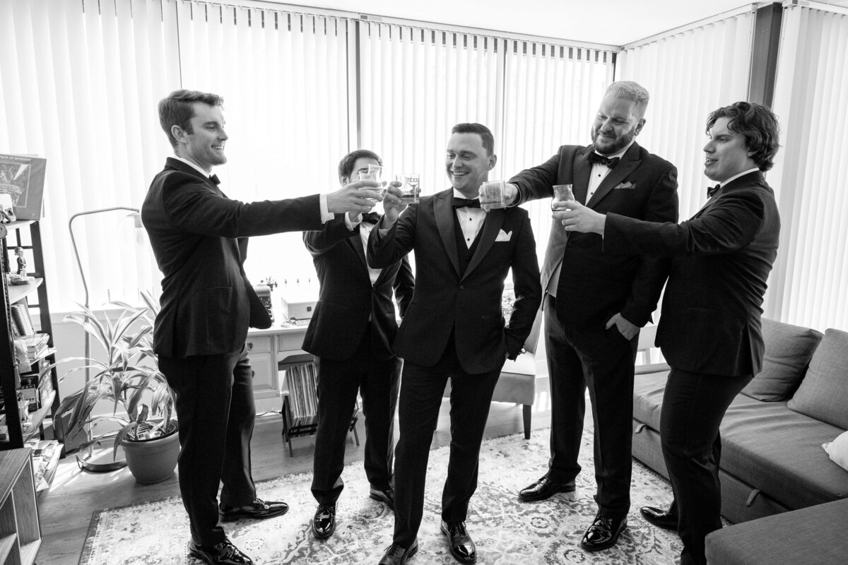 Five men in tuxedos stand in a living room, raising glasses in a toast. They are smiling and appear to be celebrating.
