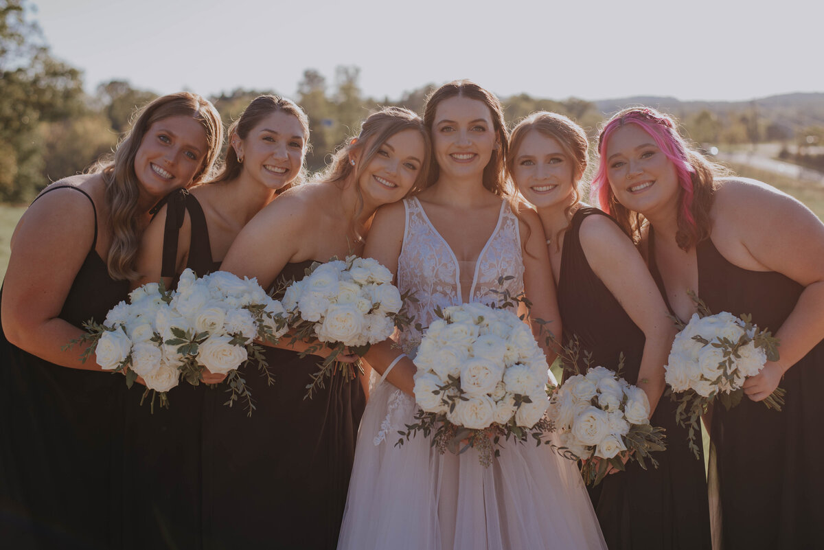 A bride in a white dress stands outdoors at a rustic wedding venue, surrounded by five bridesmaids in black dresses. They all hold bouquets of white flowers and smile toward the camera, set against a sunlit, grassy backdrop—an idyllic scene for perfect wedding packages in Virginia.