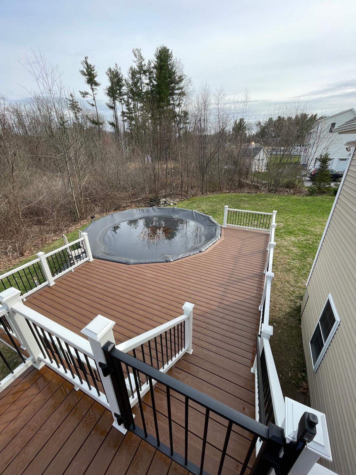 A beautiful dark brown deck with white railings and trellis stairs facing around a pool done by a Northborough Deck Contractor