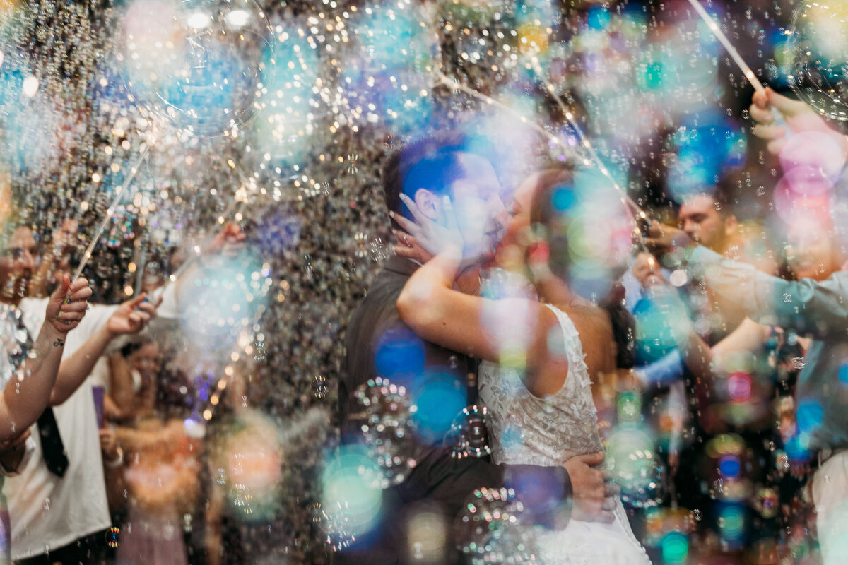 Bride and groom share a kiss during their Lake Mary Event Center wedding reception exit, surrounded by bubbles and colorful lights.